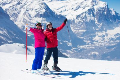Senior couple stands on a ski slope with mountains in the background.